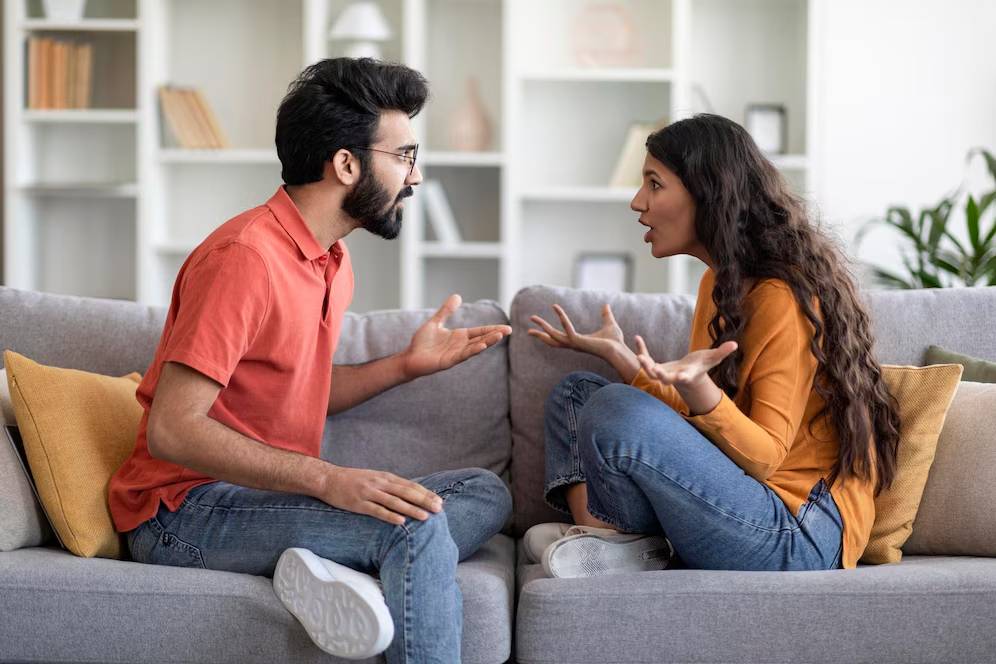 Two people having an animated conversation on a gray sofa in a living room.