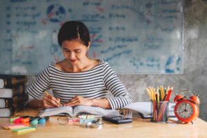 Woman studying with books, stationery, and a red clock on a wooden table, with a chalkboard in the background.