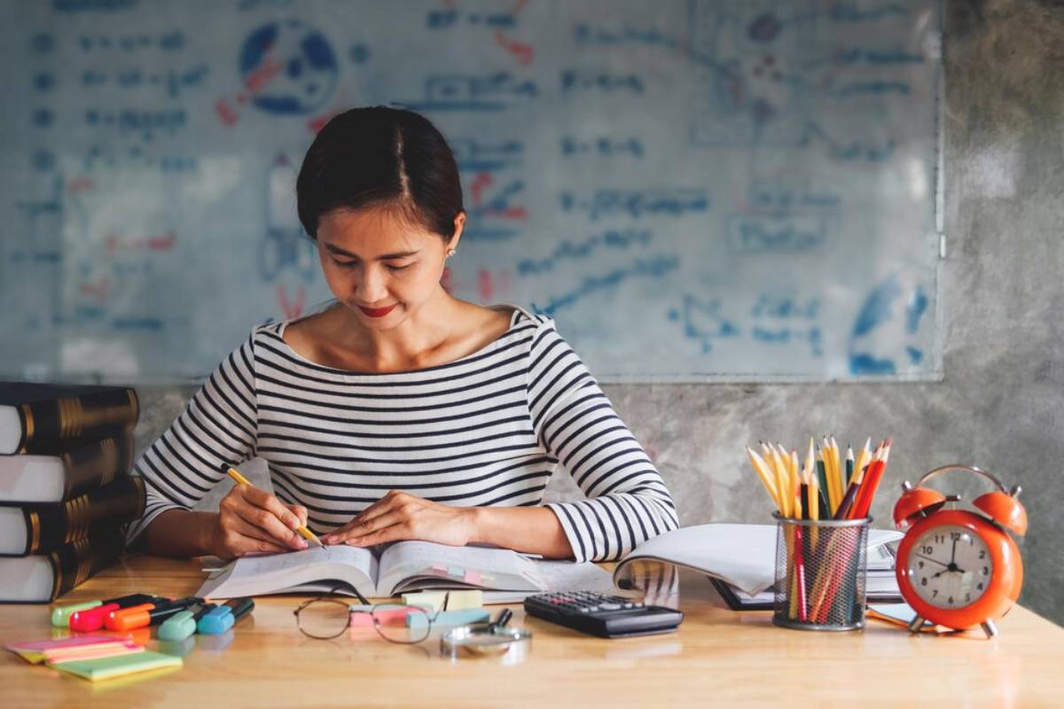 Woman studying with books, stationery, and a red clock on a wooden table, with a chalkboard in the background.