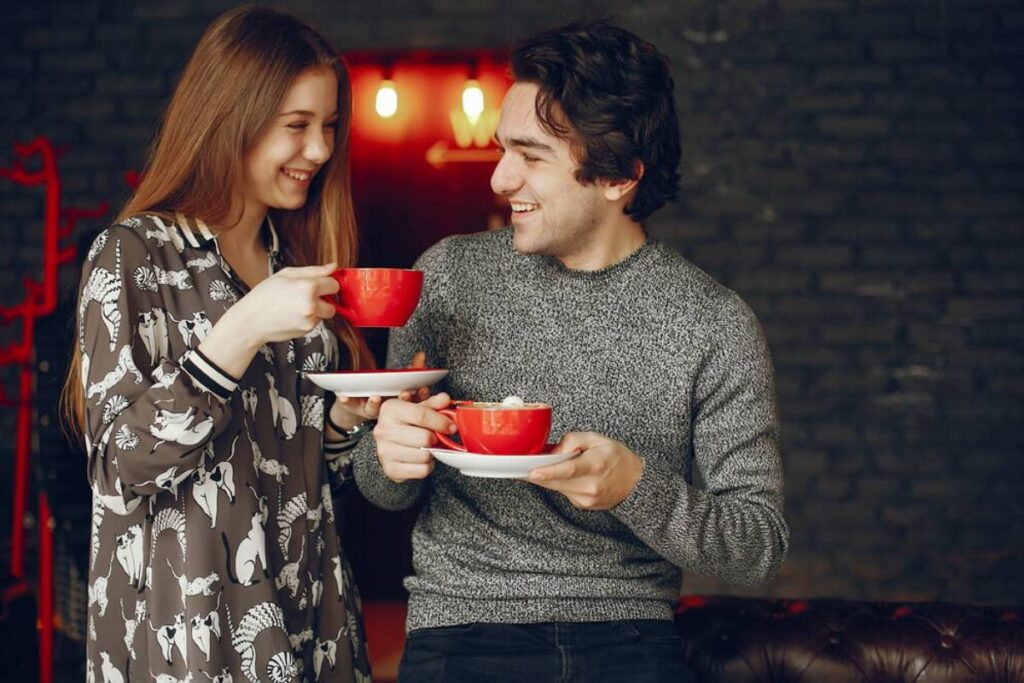 A young man and woman happily holding and clinking red coffee cups in a cozy, dimly lit room.