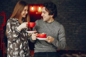 A young man and woman happily holding and clinking red coffee cups in a cozy, dimly lit room.