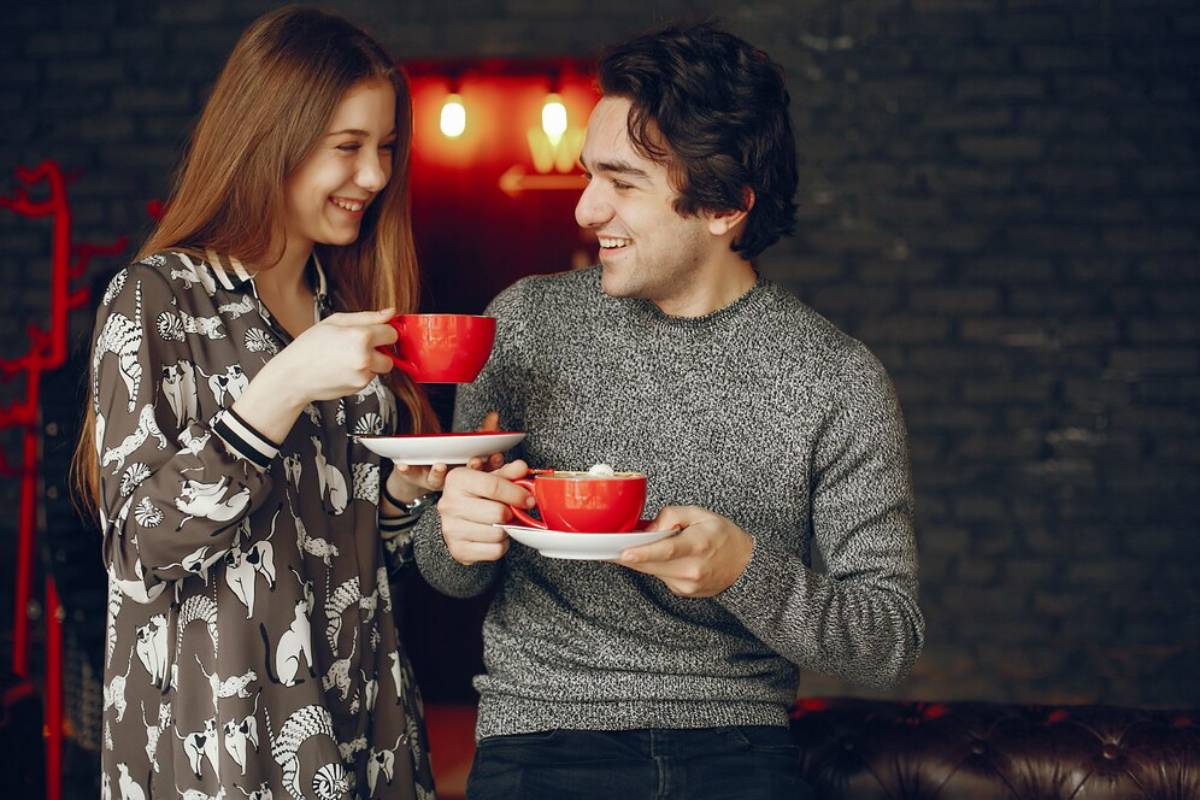 A young man and woman happily holding and clinking red coffee cups in a cozy, dimly lit room.