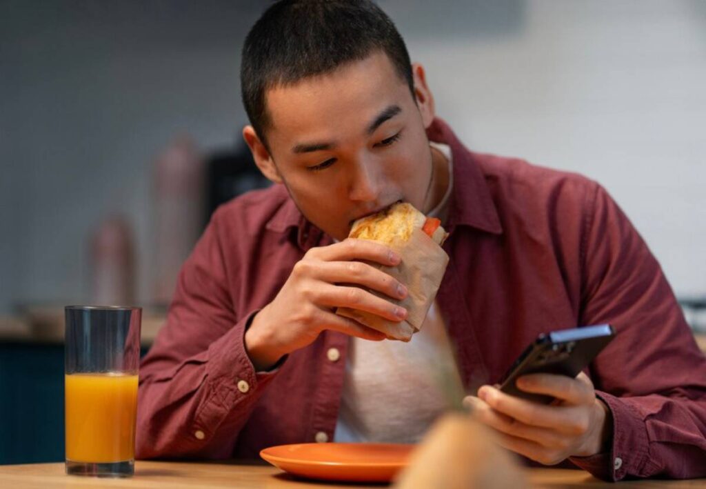 A person sits at a table, holding a wrapped food item in one hand and a smartphone in the other, with a glass of orange juice nearby.