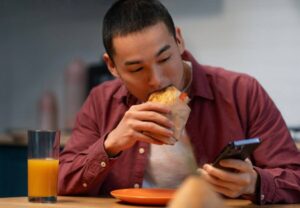 A person sits at a table, holding a wrapped food item in one hand and a smartphone in the other, with a glass of orange juice nearby.