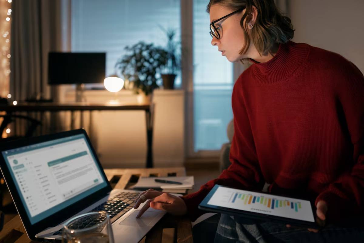 Young woman with glasses working on laptop and tablet in a dimly lit room at night.