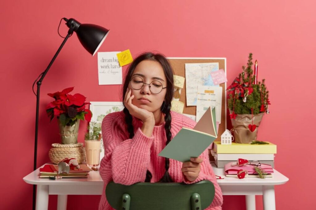 Young woman in glasses and pink sweater looks bored while holding a book, sitting at a desk with festive decorations and notes.