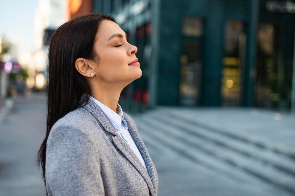 Woman relaxing with eyes closed on a city street, wearing a gray coat and a white shirt.