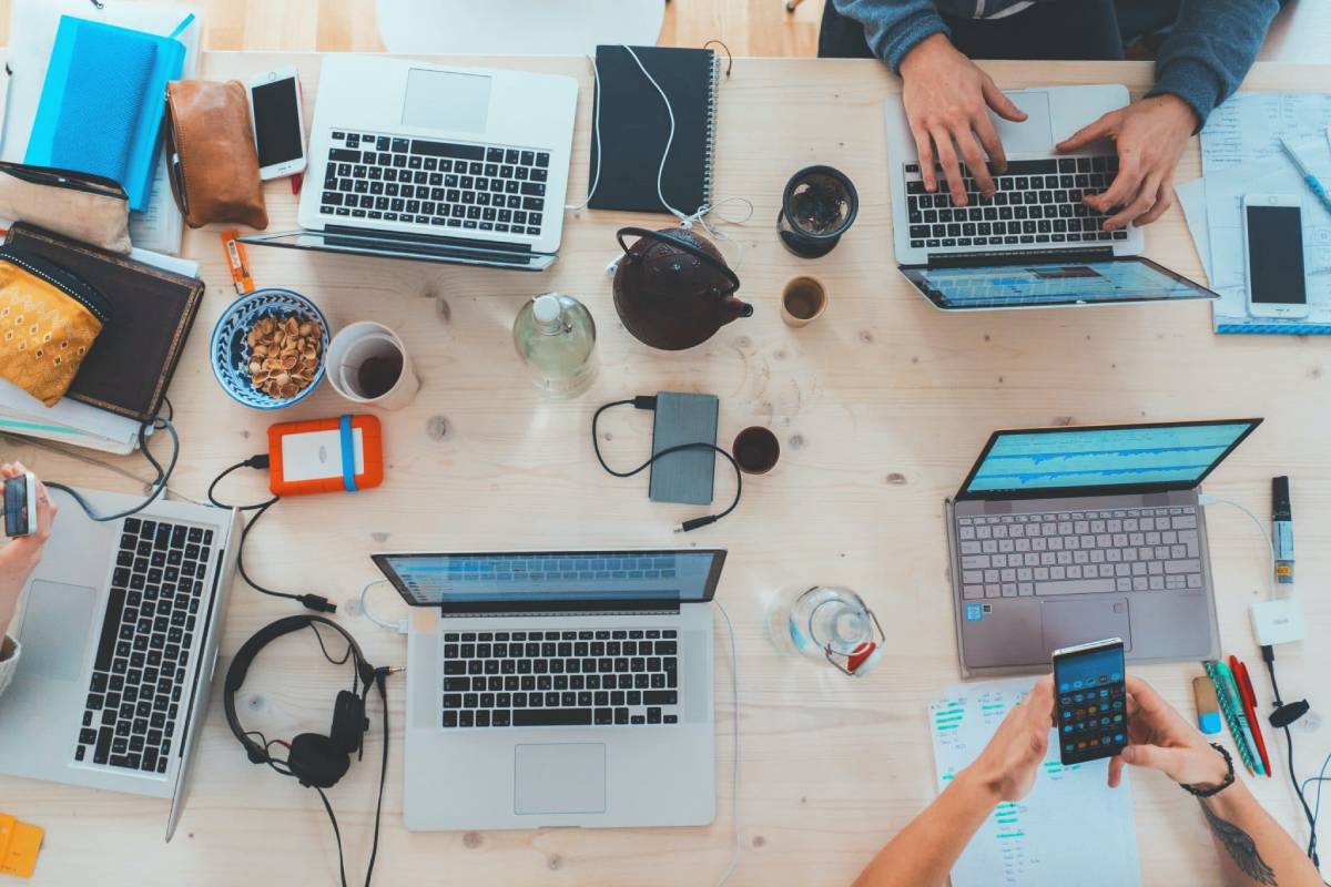 Overhead view of a busy workspace with multiple laptops, notes, and electronic devices on a wooden table.