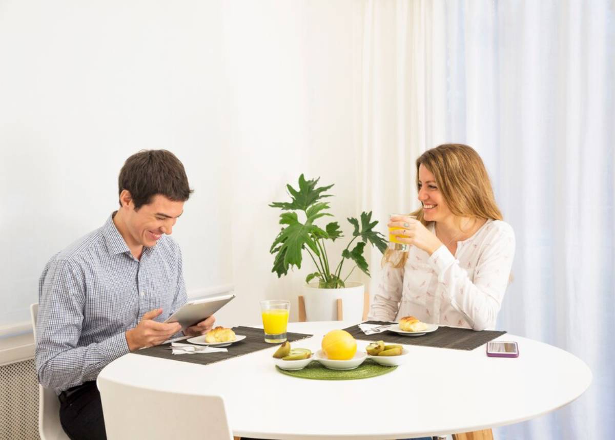 A man and woman enjoying breakfast together in a bright dining room, with the man smiling at a tablet.