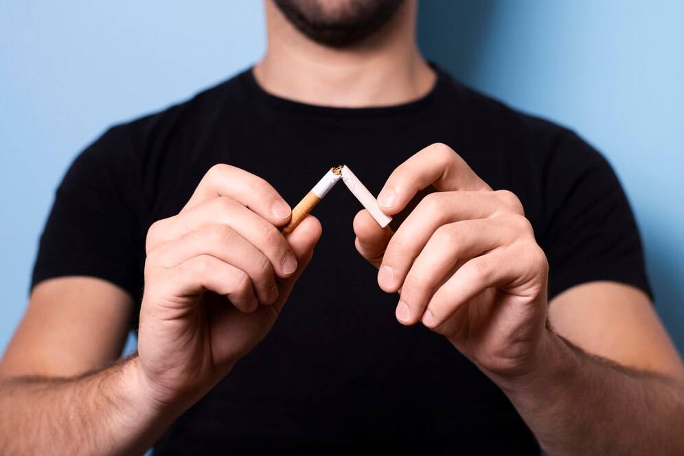 A person in a black t-shirt breaks a cigarette in half, symbolizing the act of quitting smoking. Background is light blue.