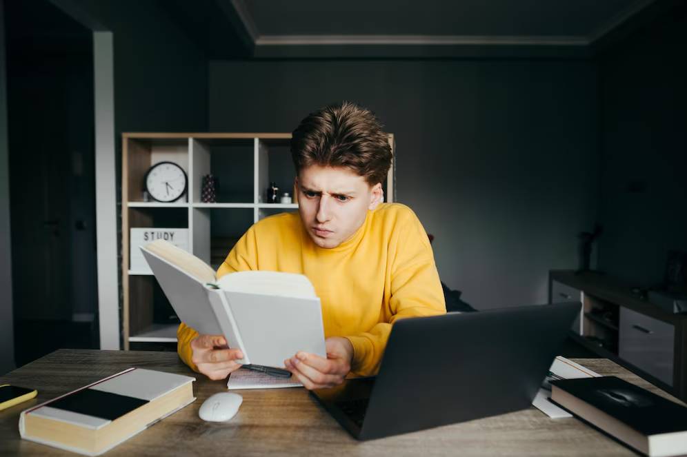Young man in a yellow sweater looking confused while reading a book and using a laptop at a desk.