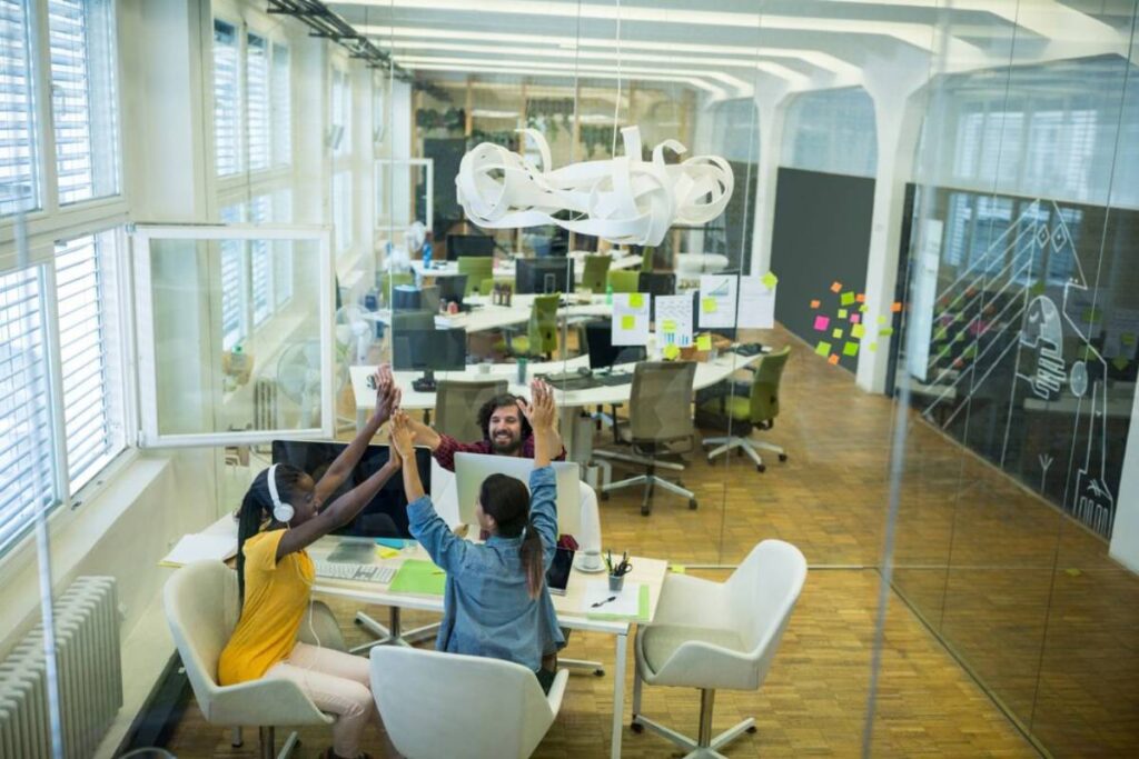 Three colleagues high-five happily in a bright, modern office with desks and paper sculptures.