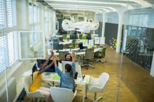 Three colleagues high-five happily in a bright, modern office with desks and paper sculptures.