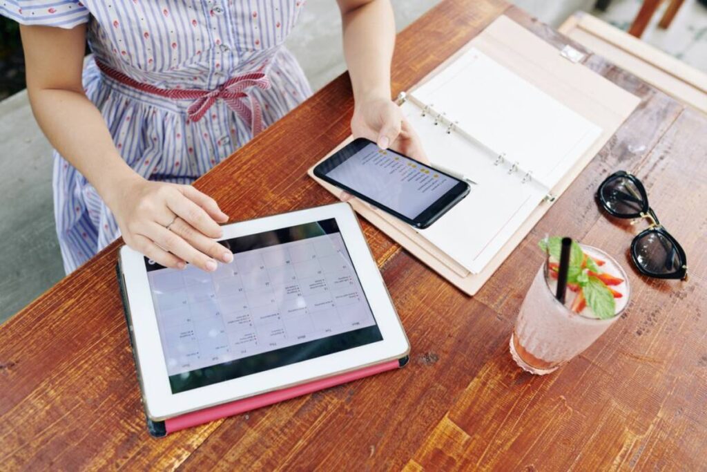 Woman at a table using a tablet and smartphone with open planner and a drink beside her.