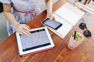 Woman at a table using a tablet and smartphone with open planner and a drink beside her.