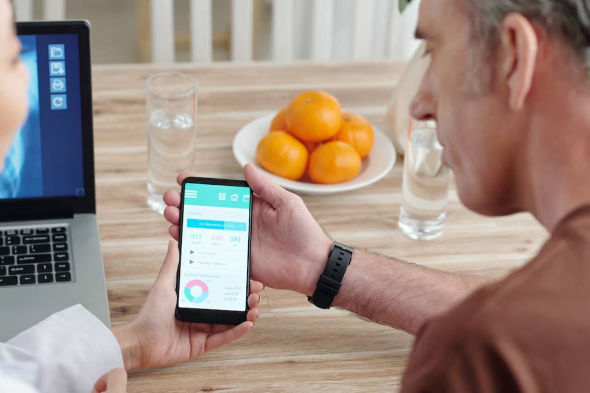 A man checks health data on his smartphone at a dining table, while another person uses a laptop nearby.