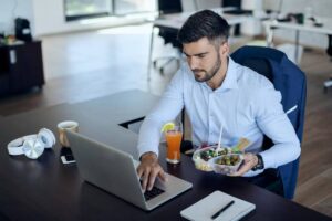 Man eating salad and working on laptop in modern office.