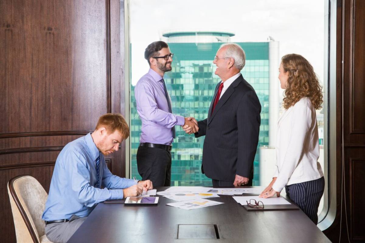 Two men shaking hands at a boardroom table while colleagues watch and work around them.