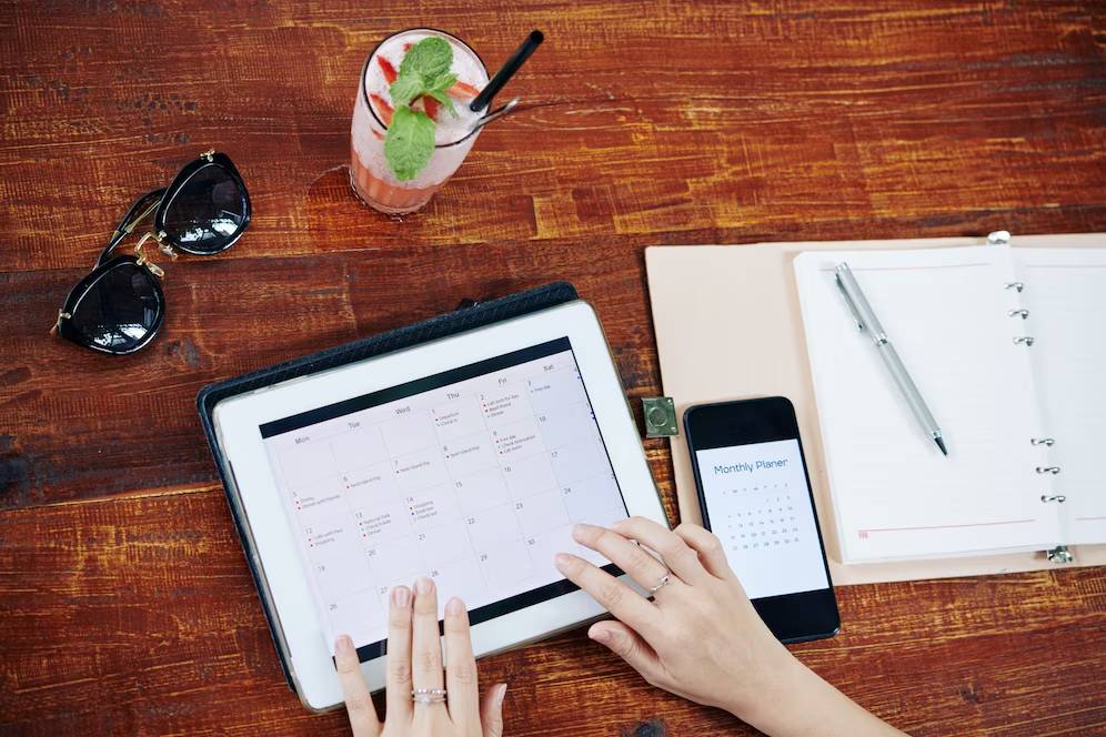A person managing a digital calendar on a tablet, with a planner, phone, cocktail, and sunglasses on a wooden table.
