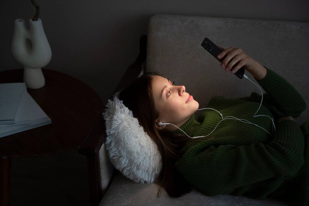 Young woman lying on a couch listening to music through earphones while holding a smartphone in a dimly lit room.