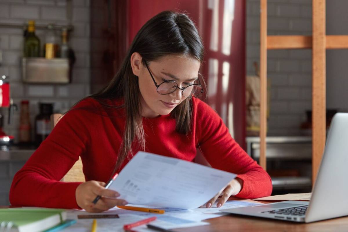 Woman in a red sweater reviews documents at a kitchen table with a laptop nearby.