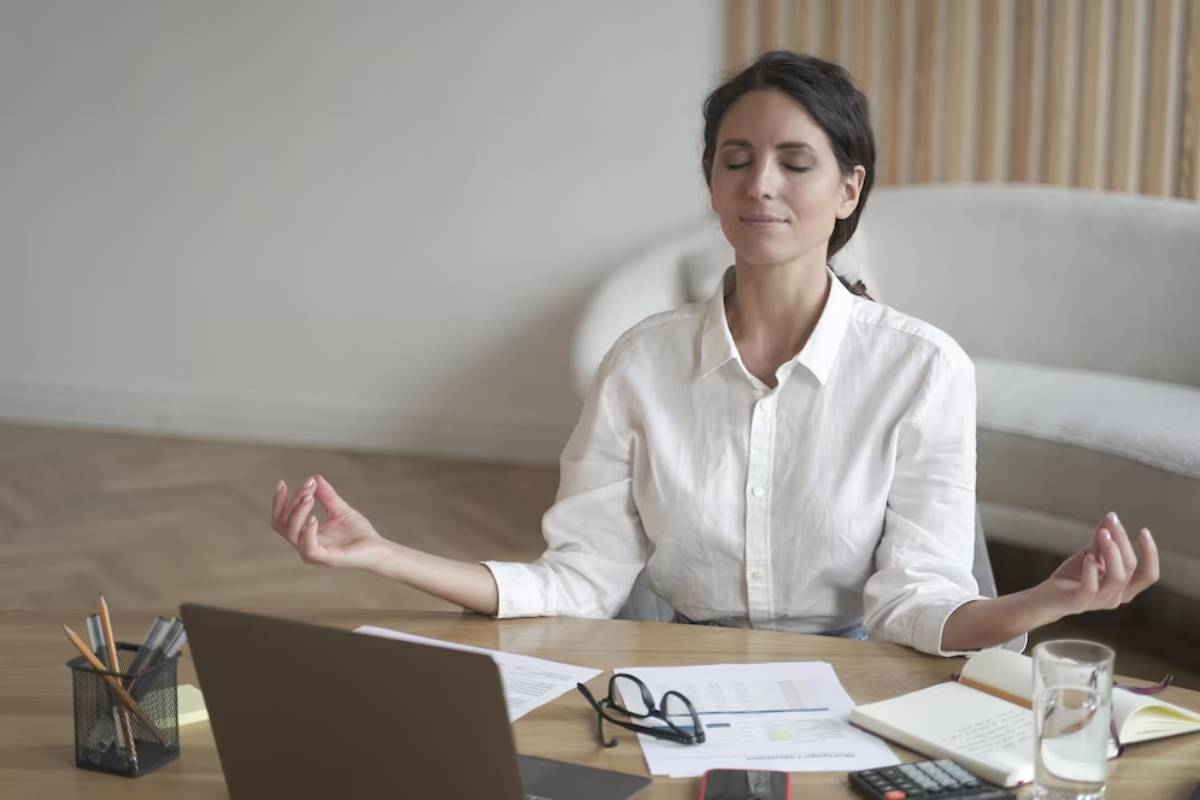 A woman meditating at a desk with a laptop, papers, and office supplies in a bright room.