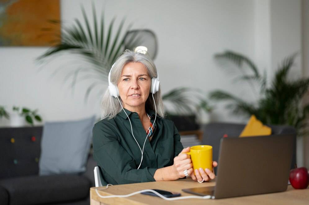 Mature woman with headphones using laptop and holding a yellow mug in a modern living room.