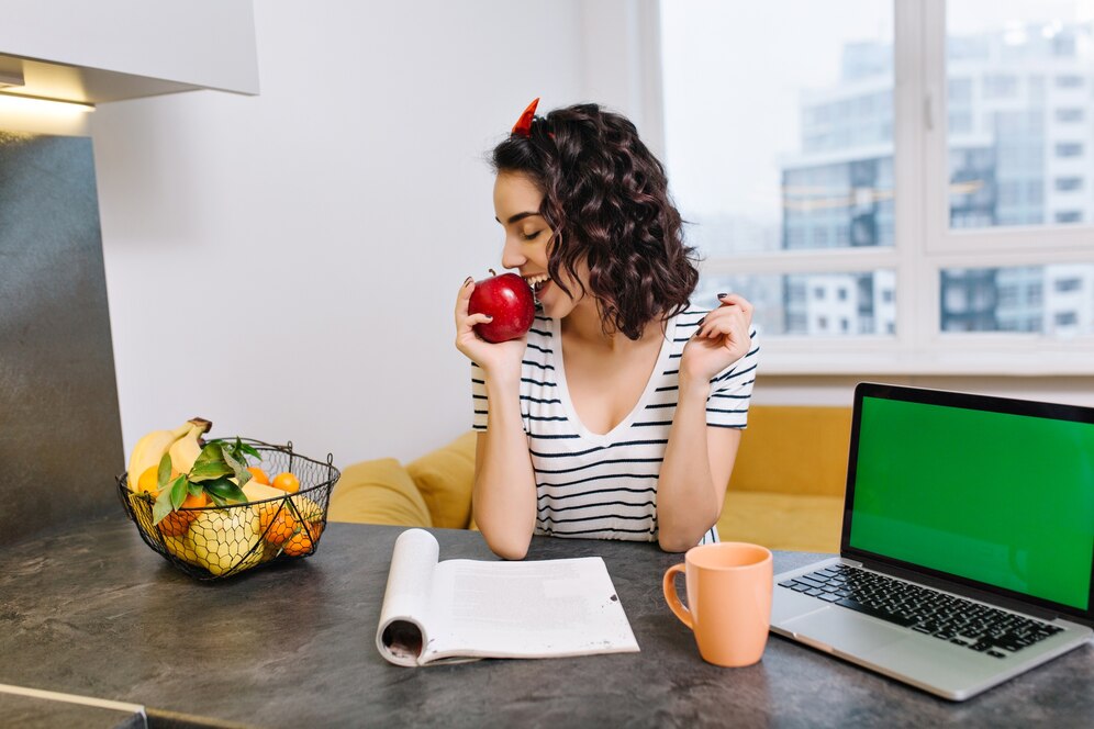 Woman eats an apple while studying with an open laptop and notebook at a kitchen table.
