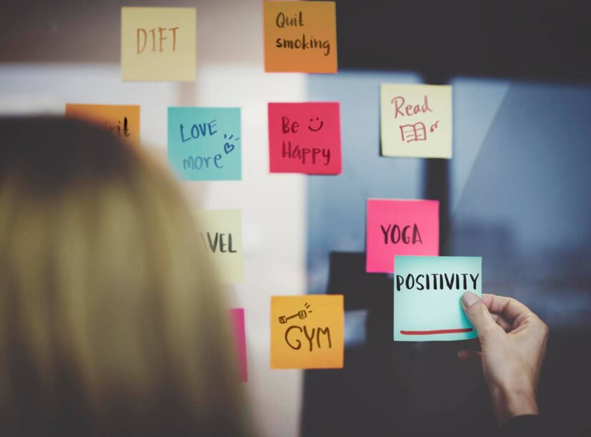 Woman placing a sticky note labeled POSITIVITY on a board full of motivational reminders.
