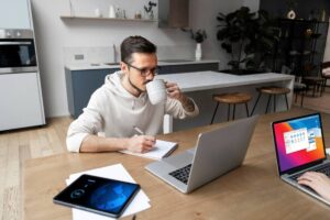 Man working at a home office with two laptops, taking notes and drinking from a mug.