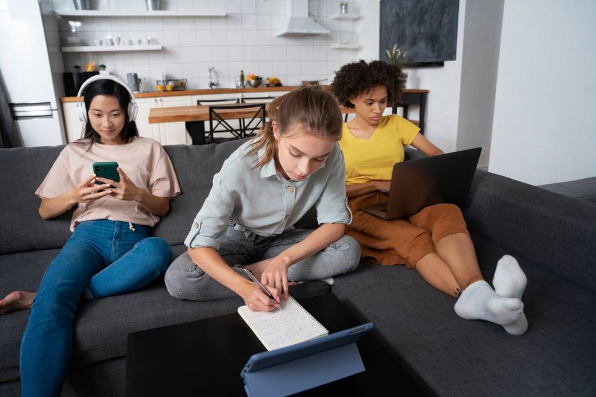 Three young people using devices on a couch; one with headphones on a phone, another on a laptop, and one writing notes.