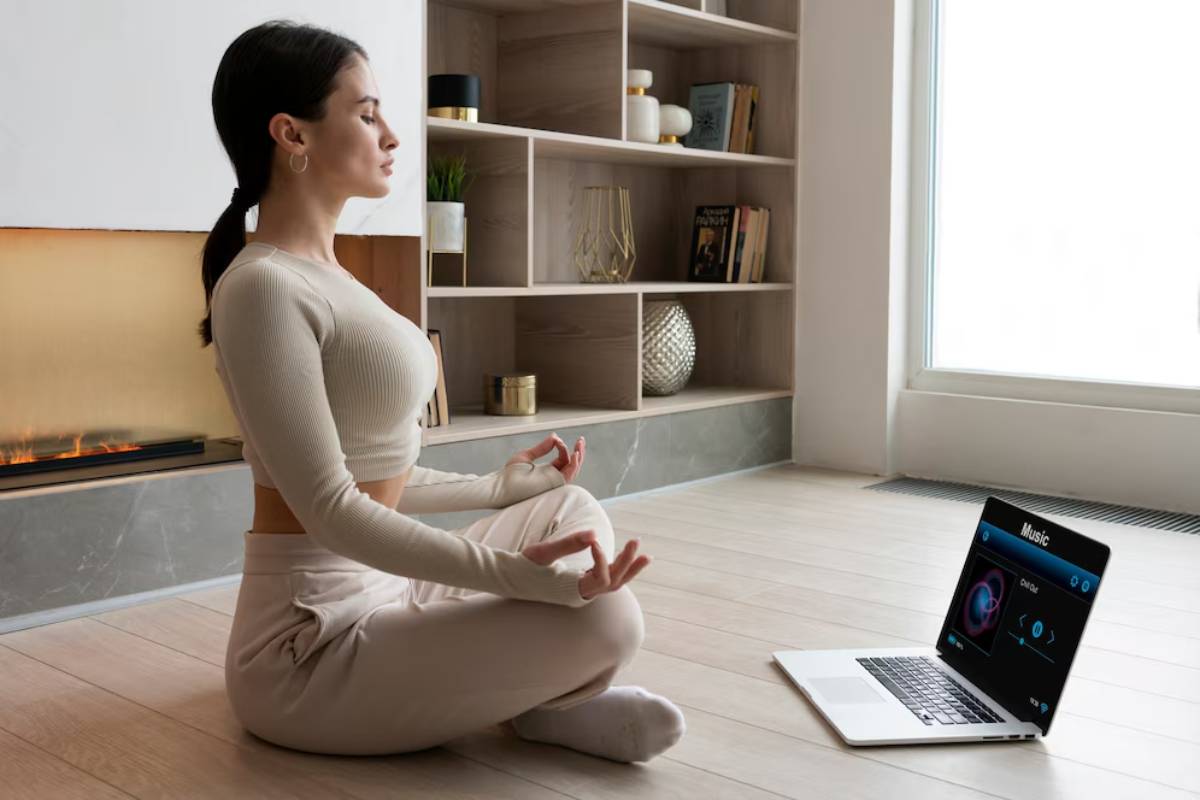 Woman meditating in a living room with a laptop showing a music app next to her.