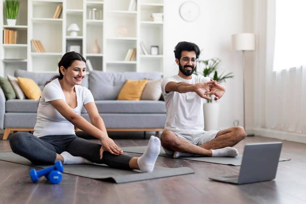 A man and woman smiling and stretching on yoga mats in a living room, following a workout video on a laptop.