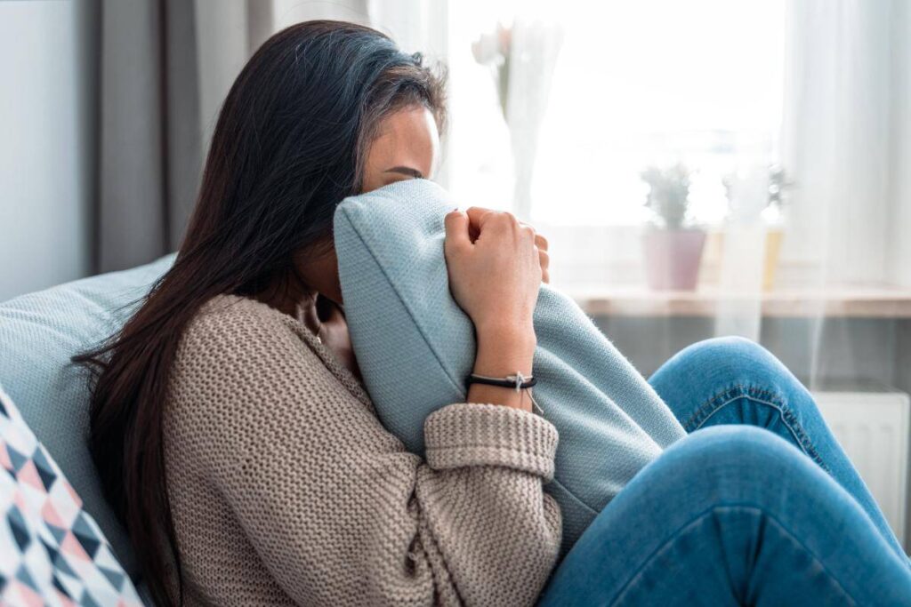 Woman sitting on a sofa, hiding her face in a pillow, looking distressed.