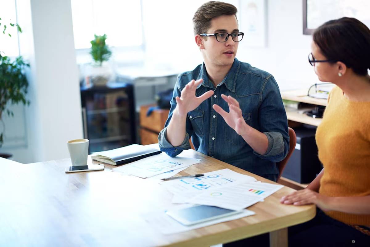 A man in glasses explaining something with hand gestures to a woman at an office desk with papers and a coffee cup.