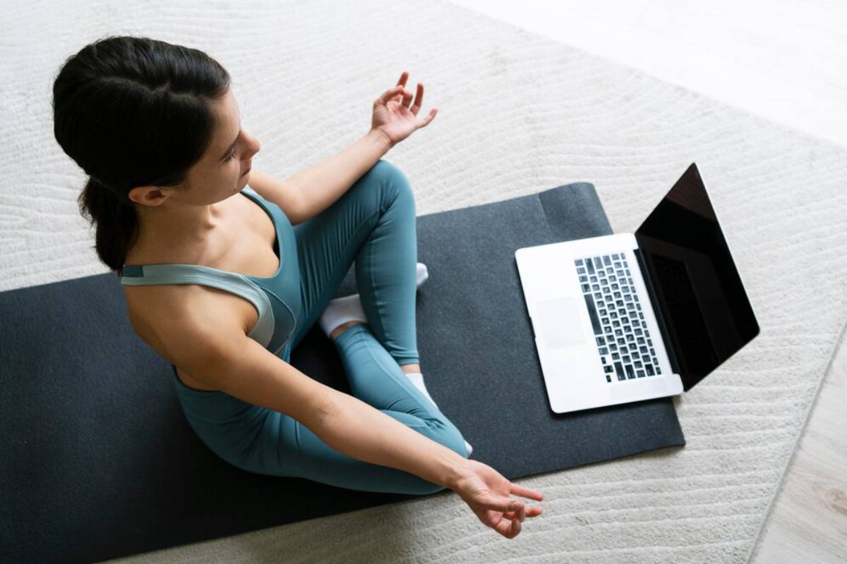 A person in yoga attire practices meditation on a mat, with a laptop nearby, against a light, serene background.