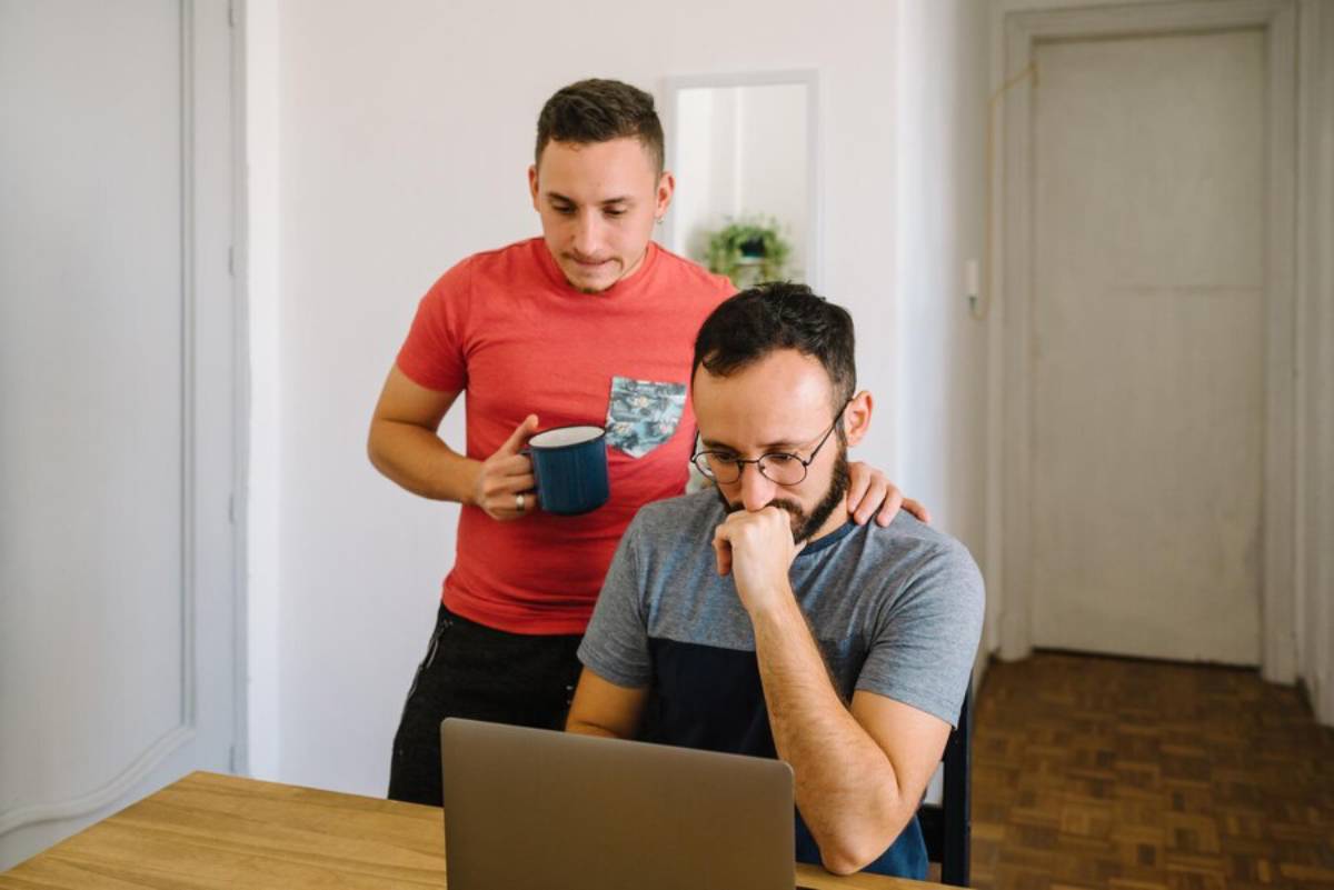 Two men in a home office, one focused on the laptop, the other holding a coffee mug and looking over his shoulder.