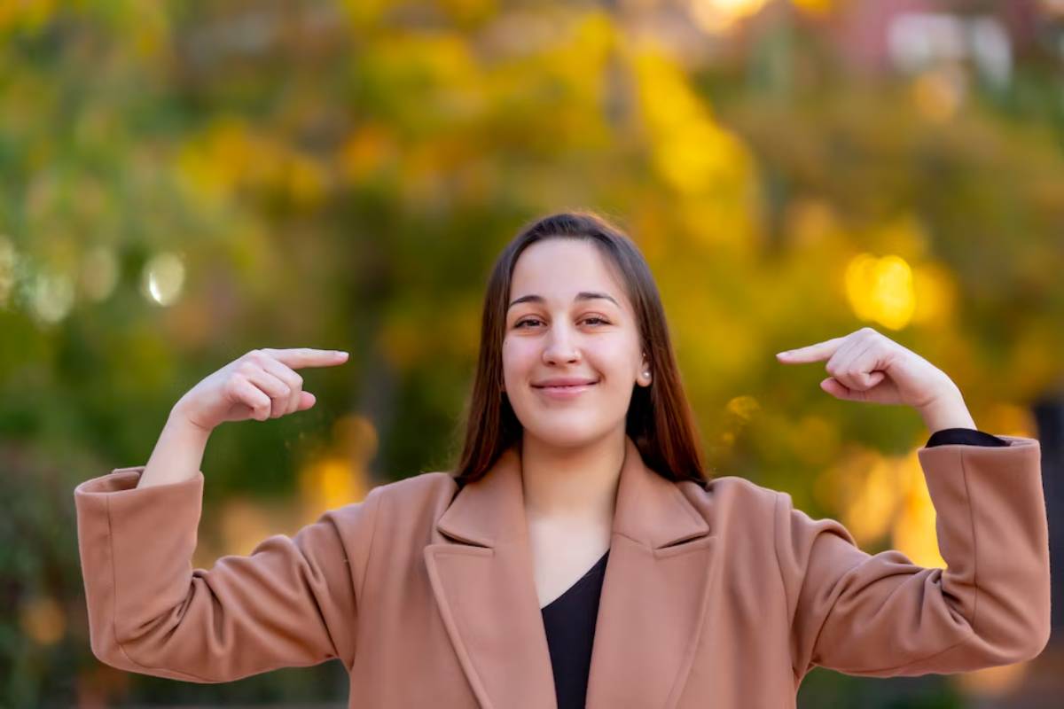 Young woman in a tan coat pointing to herself with a smile, standing before autumn foliage.