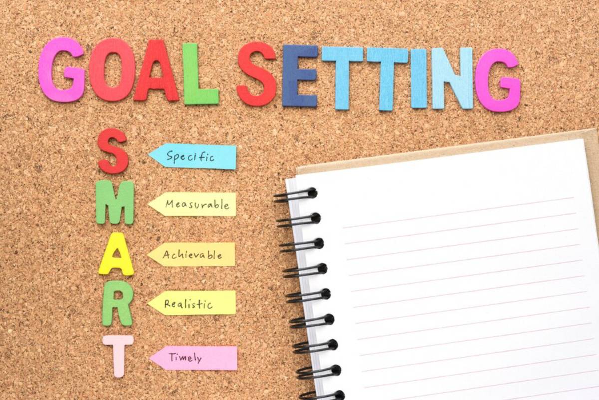 Multicolored GOAL SETTING letters and a SMART goals acronym on notes pinned to a cork board beside a blank notebook.