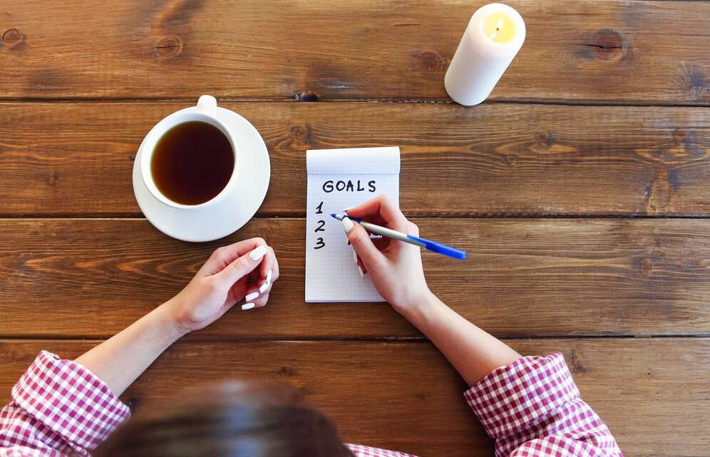 Person writing a goals list on a notepad next to a cup of tea and a candle on a wooden table.