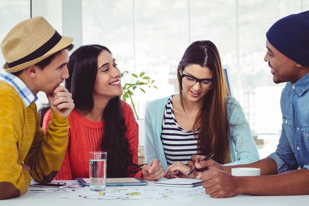 Four diverse young adults engaged in a discussion with papers and digital devices on a table in a bright office setting.