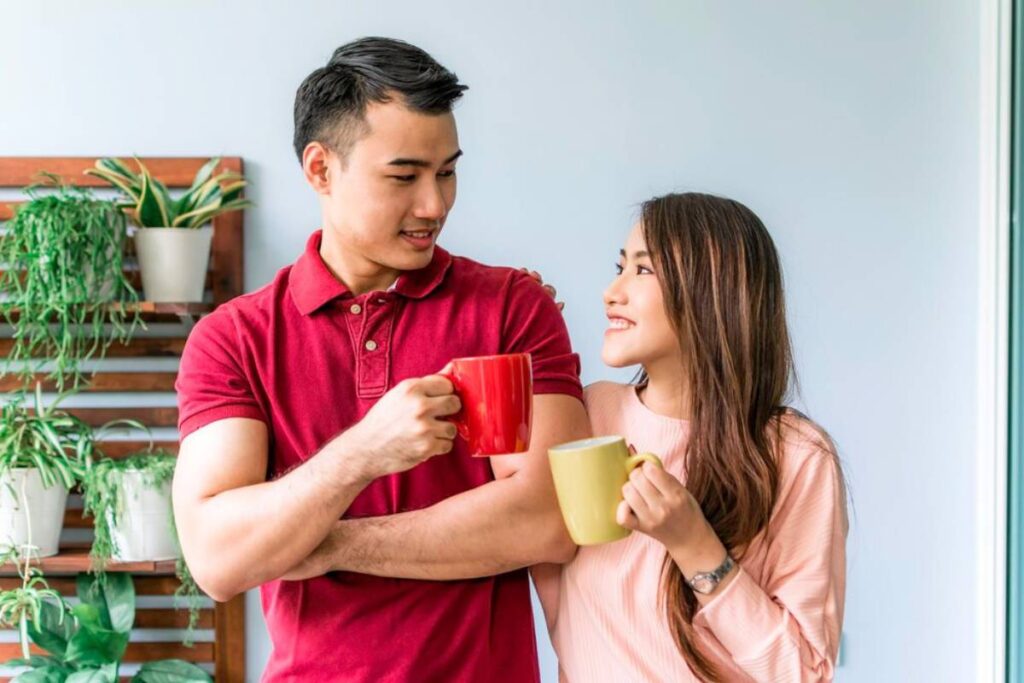 Man and woman smiling and clinking coffee mugs indoors, surrounded by houseplants.