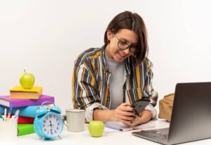 Young woman smiling at her phone, seated at a desk with a laptop, books, and a clock.