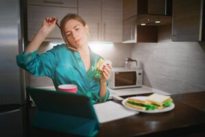 Woman in a teal robe eating a sandwich in a modern kitchen, looking tired or contemplative.