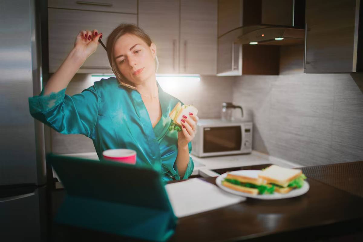 Woman in a teal robe eating a sandwich in a modern kitchen, looking tired or contemplative.