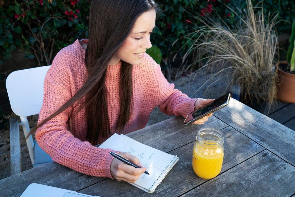 A woman in a pink sweater writes in a notebook and uses a smartphone at an outdoor table with a jar of juice.