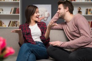 A young woman and man engaging in a lively conversation on a sofa, surrounded by bookshelves.