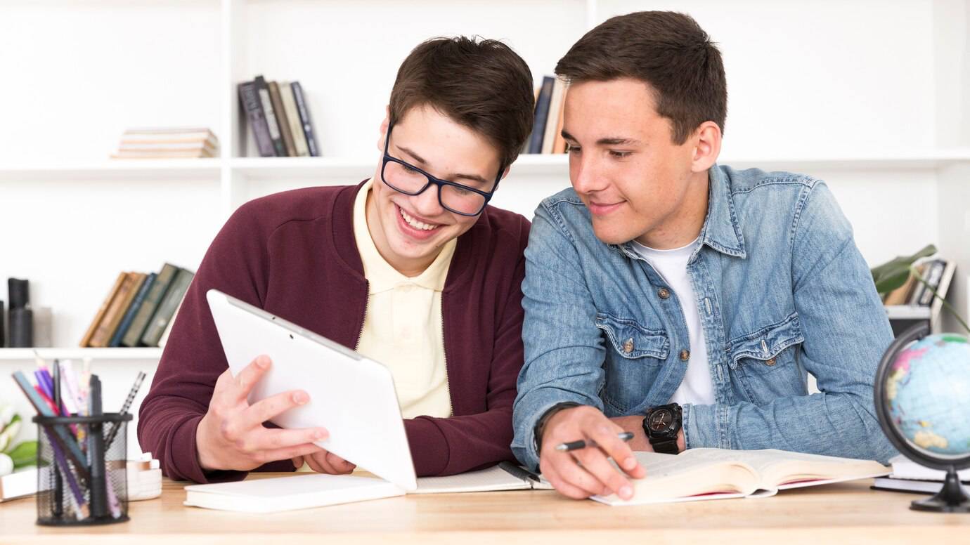 Two young men are studying together at a wooden table, using a tablet and books, surrounded by shelves of books and a globe.