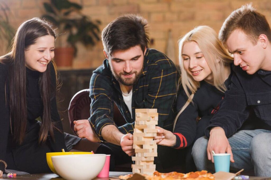 A group of friends focus on a game of Jenga on the floor, surrounded by snacks and drinks in a cozy indoor setting.