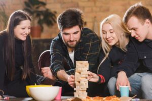 A group of friends focus on a game of Jenga on the floor, surrounded by snacks and drinks in a cozy indoor setting.